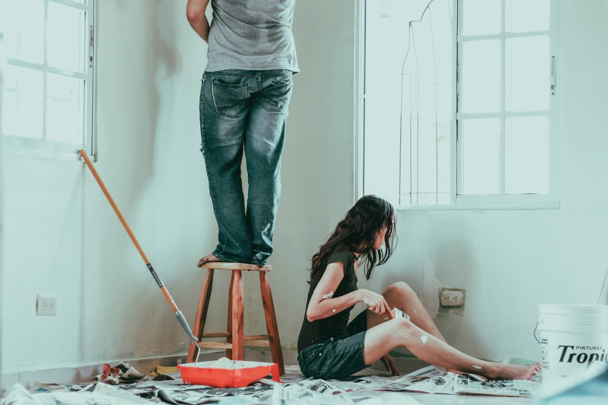 Twee personen voeren renovatiewerkzaamheden uit in een kamer met verfgereedschap en beschermingspapier.